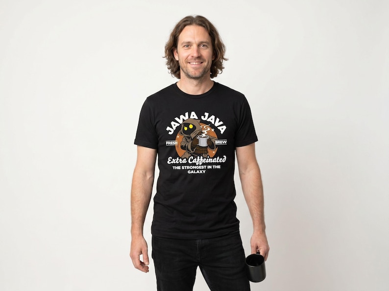 May include: Black t-shirt with a Jawa Java graphic. The design features a cartoon Jawa character holding a coffee cup, with text that reads "Extra Caffeinated: The Strongest in the Galaxy." The shirt is worn by a person holding a black mug.