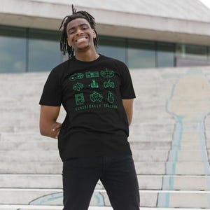 May include: A black short-sleeved t-shirt featuring a green pixelated graphic of various vintage video game controllers and a handheld console, with the text "CLASSICALLY TRAINED" printed in green capital letters underneath the icons.