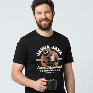 May include: Black t-shirt featuring the text "JAWA JAVA" and an illustration of a Jawa with a coffee mug. The shirt also displays "Extra Caffeinated" and "The Strongest in the Galaxy". A black coffee mug is held in the hand.