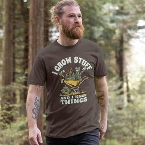 May include: A dark brown short-sleeve t-shirt featuring a graphic of a yellow wheelbarrow filled with garden tools and plants. The text "I GROW STUFF AND I KNOW THINGS" is printed in a cream-coloured font above and below the wheelbarrow illustration.