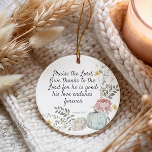 Puede incluir: Adorno navideño redondo de cerámica blanca con un lazo dorado para colgar. Presenta ilustraciones florales y el texto "Praise the Lord, Give thanks to the Lord for he is good; his love endures forever" y "Psalm 106:1".