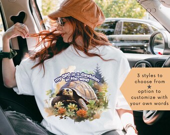 Op de afbeelding: Wit T-shirt met een schildpad afbeelding en de tekst "my GPS is a dandelion in the wind". Extra tekst luidt "3 styles to choose from" en "option to customize with your own words."