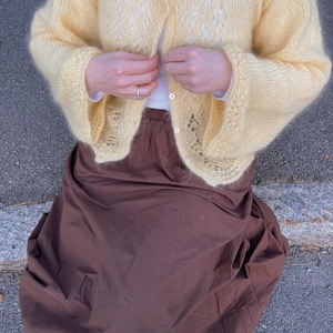 Könnte beinhalten: Eine hellgelbe, langärmelige Strickjacke mit Knöpfen und einem zarten, durchbrochenen Spitzenmuster auf den Vorderteilen. Die Strickjacke wird über einem weißen Oberteil getragen und mit einem langen, einfarbigen dunkelbraunen Rock kombiniert.