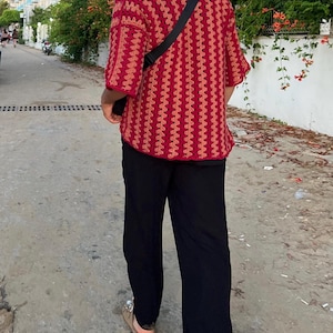 Peut inclure: Une chemise en crochet rouge et beige à motif vertical est associée à un pantalon noir. La chemise a des manches courtes et un sac à bandoulière noir est porté en travers du corps. La personne marche sur un chemin pavé.