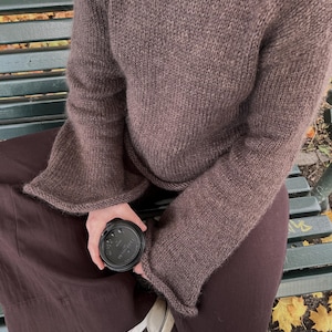 May include: A brown knit sweater with flared sleeves. The person is wearing brown pants and white sneakers with black stripes. A black coffee cup is held in the person's hand. The person is sitting on a green bench with yellow leaves on the ground.