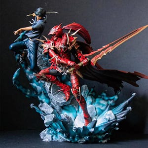 Può includere: Statuetta dettagliata raffigurante due personaggi in pose dinamiche. Uno è vestito con un'armatura rossa e brandisce una grande spada, l'altro indossa abiti blu e una spada. La base presenta accenti blu e grigi, creando una scena drammatica.