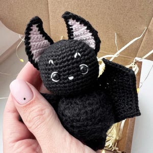 May include: A black crocheted bat with pink ears and white eyes. The bat is holding its wings out to the sides.