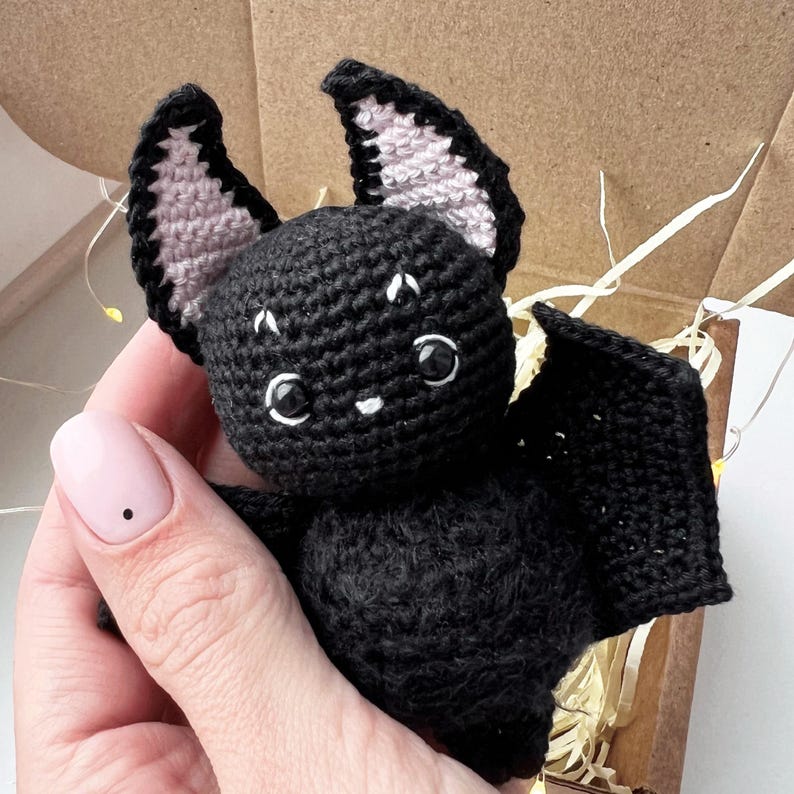 May include: A black crocheted bat with pink ears and white eyes. The bat is holding its wings out to the sides.