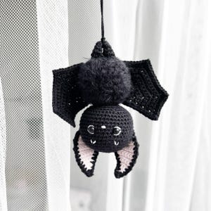 May include: A black crocheted bat with pink ears hanging from a white curtain. The bat has a fuzzy head and black eyes.