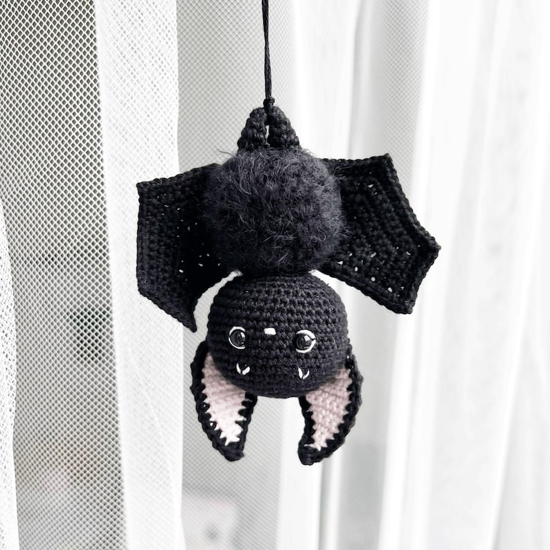May include: A black crocheted bat with pink ears hanging from a white curtain. The bat has a fuzzy head and black eyes.