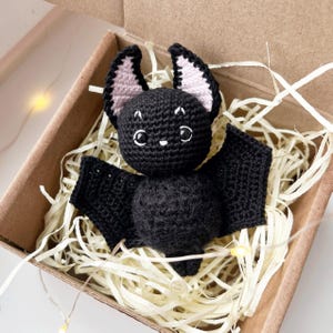 May include: A black crocheted bat with pink ears, sitting in a brown box filled with shredded paper. The bat has large, black eyes and a small, black nose.