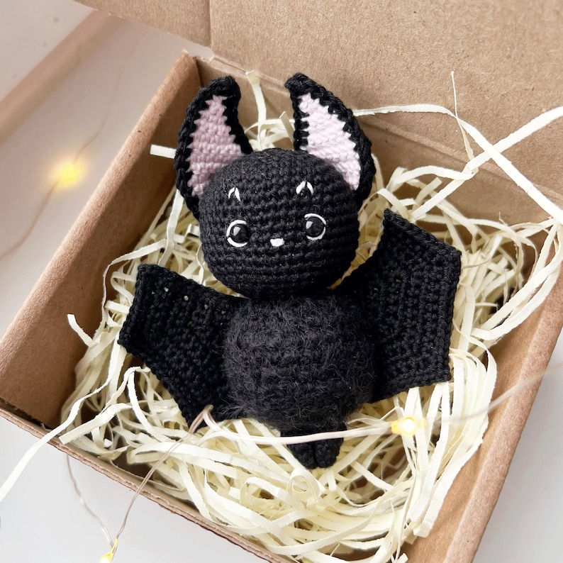 May include: A black crocheted bat with pink ears, sitting in a brown box filled with shredded paper. The bat has large, black eyes and a small, black nose.