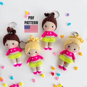 Op de afbeelding: Vier handgemaakte gehaakte poppen sleutelhangers. Elke pop heeft een andere haarkleur: bruin, blond en twee met bruin haar. Ze dragen roze tops en groene rokken. De sleutelhangers liggen op een wit oppervlak met kleurrijke hart- en stervormige decoraties. De afbeelding bevat de tekst "PDF PATTERN" en een Amerikaanse vlag.