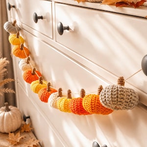 May include: A handmade crochet pumpkin garland in shades of orange, yellow, and cream, strung on a natural twine. The garland is displayed against a white dresser, creating a festive autumn decoration.