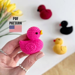 Puede incluir: Un patrón de crochet para hacer un pato rosa con un pico amarillo. El patrón está disponible para descargar en PDF. La imagen también muestra un pato negro, un pato amarillo y un pato rojo.
