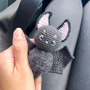 May include: A small crocheted bat toy with gray fur, pink ears, and black eyes. The bat is being held by a hand.