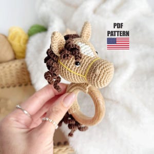 以下が含まれることがあります： 木製の輪が付いた、編みぐるみのおもちゃの馬。馬は茶色と白で、たてがみと尻尾は黄色です。赤ちゃんや幼児に最適なおもちゃです。