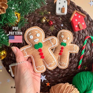 Puede incluir: Dos adornos de hombre de jengibre de ganchillo con detalles de glaseado blanco, lazos verdes y acentos de botones rojos. Los adornos se exhiben sobre una estera tejida con decoraciones navideñas. El texto "PDF PATTERN" es visible.