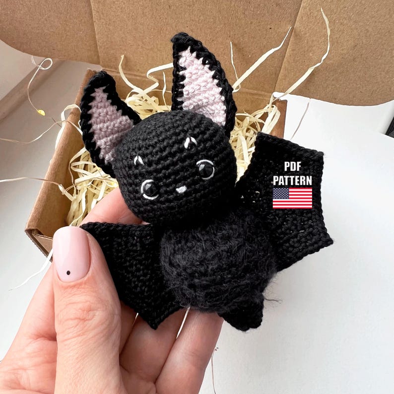 May include: A crocheted black bat with pink ears. The bat has large, round eyes and a small, white mouth. The bat is sitting in a box filled with shredded paper. The text "PDF PATTERN" is visible in the image.