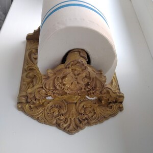 May include: An antique-style paper towel holder with a gold finish. The holder has detailed scrollwork and floral patterns. A roll of white paper towels with blue stripes is in the holder.