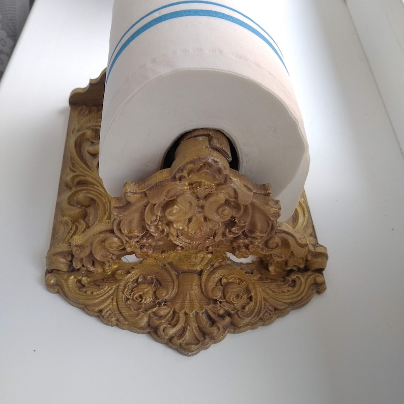 May include: An antique-style paper towel holder with a gold finish. The holder has detailed scrollwork and floral patterns. A roll of white paper towels with blue stripes is in the holder.