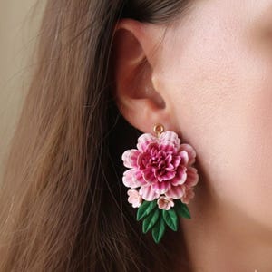 以下が含まれることがあります： 大きなピンクと白の層状の花、小さなピンクの花、緑の葉が特徴のフローラルイヤリング。イヤリングは金色のフックに取り付けられています。