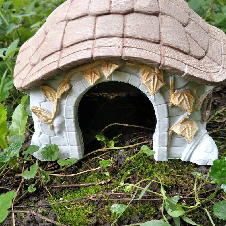 May include: A miniature fairy house with a light brown tiled roof and a white stone facade. The arched doorway is framed by decorative golden leaf accents. The house is surrounded by green foliage and moss.