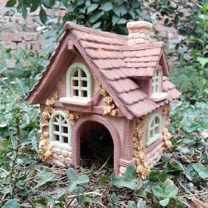 May include: A miniature cottage figurine featuring a terracotta-colored roof with a chimney, light-colored walls with arched windows, and decorative relief carvings of climbing vines. The structure has an open arched doorway at the front.