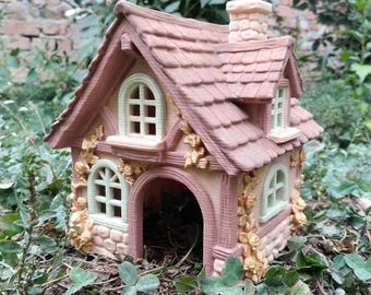 May include: A miniature cottage figurine featuring a terracotta-colored roof with a chimney, light-colored walls with arched windows, and decorative relief carvings of climbing vines. The structure has an open arched doorway at the front.