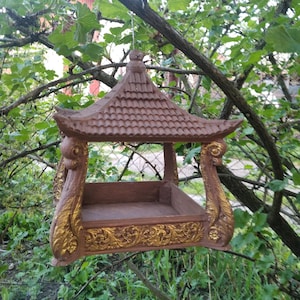 May include: A brown wooden bird feeder shaped like a pagoda with a gold trim. The feeder has a flat, rectangular base and a roof with a pointed top.