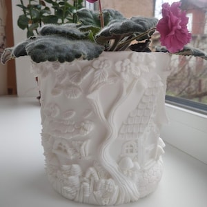 May include: A white ceramic plant pot with a detailed fairy tale design. The pot features embossed images of houses, trees, and foliage. A flowering plant with green leaves and pink flowers sits inside the pot, adding a splash of colour.