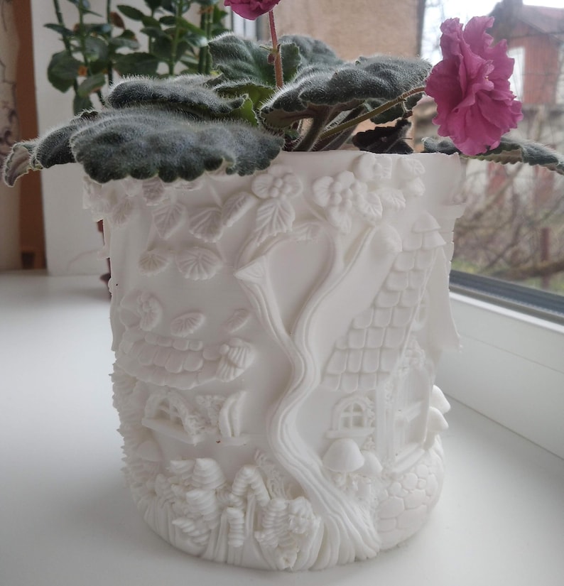 May include: A white ceramic plant pot with a detailed fairy tale design. The pot features embossed images of houses, trees, and foliage. A flowering plant with green leaves and pink flowers sits inside the pot, adding a splash of colour.