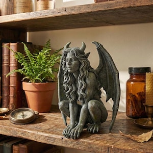 May include: A gray gargoyle statue with horns and wings, perched on a wooden shelf. The statue has long, flowing hair and is in a crouching position. A potted fern, a compass, and other decorative items are also on the shelf.