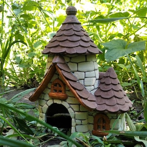 May include: A miniature fairy house with a stone effect exterior and a brown, tiled roof. The house has arched doorways and windows with orange detailing. The structure is set amongst green foliage, creating a charming garden ornament.