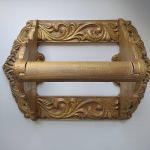 May include: A gold-painted toilet roll holder with an elaborate, decorative design. The holder has a rectangular frame with detailed scrollwork and a central roller. The style is vintage, suitable for a bathroom setting.