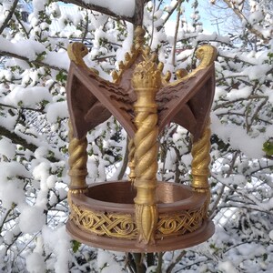 May include: A bird feeder with a decorative, ornate design. The feeder is made of brown wood with gold accents and has a round, open top. The feeder is hanging from a tree branch in a snowy winter scene.