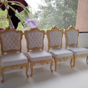 Puede incluir: Cuatro sillas con marcos de madera dorada ornamentados y tapicería blanca capitoné. Cada silla presenta un respaldo alto con detalles tallados intrincados en los bordes y cuatro patas curvas con adornos de volutas doradas a juego.