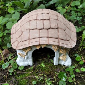 May include: A beige and gray decorative garden house with a textured roof and arched entrance. The house is made of a stone-like material and is surrounded by greenery. The roof has a tile-like pattern.