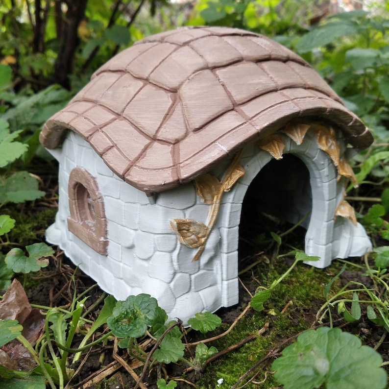 May include: A miniature fairy house with a light brown tiled roof and a white stone-like facade. The arched doorway and window are framed in brown. Decorative gold-colored leaves and vines adorn the structure, creating a whimsical garden accent.