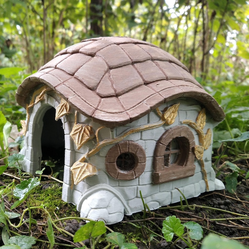 May include: A miniature fairy house with a brown tiled roof and light gray stone walls. The house features an arched doorway, round and arched windows, and decorative leaf accents. The house is set in a natural outdoor environment.