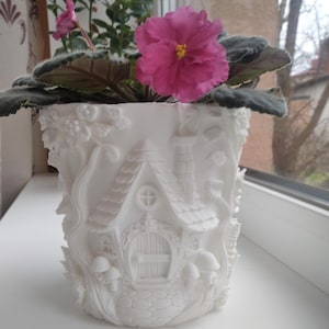 May include: A white ceramic plant pot with a detailed fairy house design. The pot features a house with a chimney, door, and windows, surrounded by sculpted mushrooms and foliage. A vibrant pink African violet plant with green leaves sits inside.