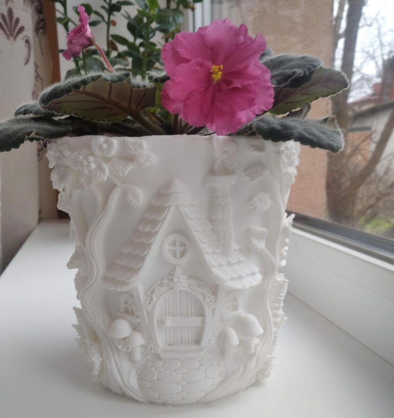May include: A white ceramic plant pot with a detailed fairy house design. The pot features a house with a chimney, door, and windows, surrounded by sculpted mushrooms and foliage. A vibrant pink African violet plant with green leaves sits inside.