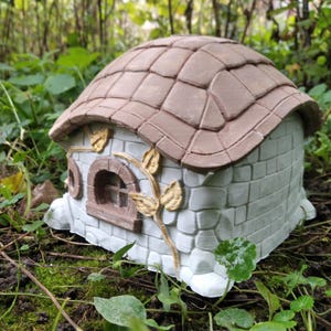 May include: A miniature fairy house with a brown shingled roof and white brick walls. The house features a small arched window and decorative gold-colored leaf accents. The whimsical structure is designed to be placed outdoors in a garden setting.