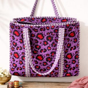 Könnte beinhalten: Eine gesteppte Tragetasche mit einem lila Leopardenmuster mit roten und schwarzen Flecken. Die Tasche hat passende lila Griffe mit weißen und grauen Streifen. Die Worte "Jaipur Block Textile" stehen am unteren Rand des Bildes.