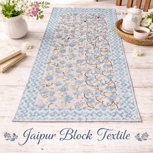 Puede incluir: Una alfombra rectangular Jaipur Block Textile con un estampado floral azul claro y blanco. La alfombra tiene un borde geométrico azul claro. El texto "Jaipur Block Textile" está en la parte inferior de la alfombra.