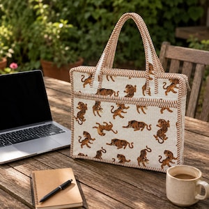 May include: A rectangular quilted laptop bag featuring a cream-colored fabric with a repeating pattern of small orange and black tigers. The bag has two matching striped handles, a zippered top closure, and a front pocket with brown striped trim.