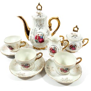 May include: A vintage porcelain tea set comprising a teapot, creamer, sugar bowl, and four cups and saucers. The set is white with gold trim and features a colorful illustration of people in period clothing. The teapot measures approximately 20 cm tall.