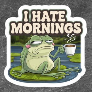 May include: A green frog with a grumpy expression sits on a lily pad and holds a cup of coffee. The text "I hate mornings" is written above the frog.