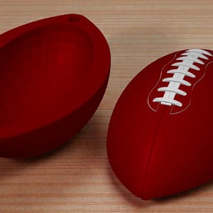 May include: A red American football with white stitching is next to a matching red, football-shaped container. The container is open, showing its inside. The objects are on a light brown, wood-grain surface.