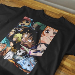 May include: Black t-shirt featuring a colorful collage of anime characters. The graphic includes various faces and figures from the anime series. The shirt is made of a dark fabric and is displayed on a wooden surface.